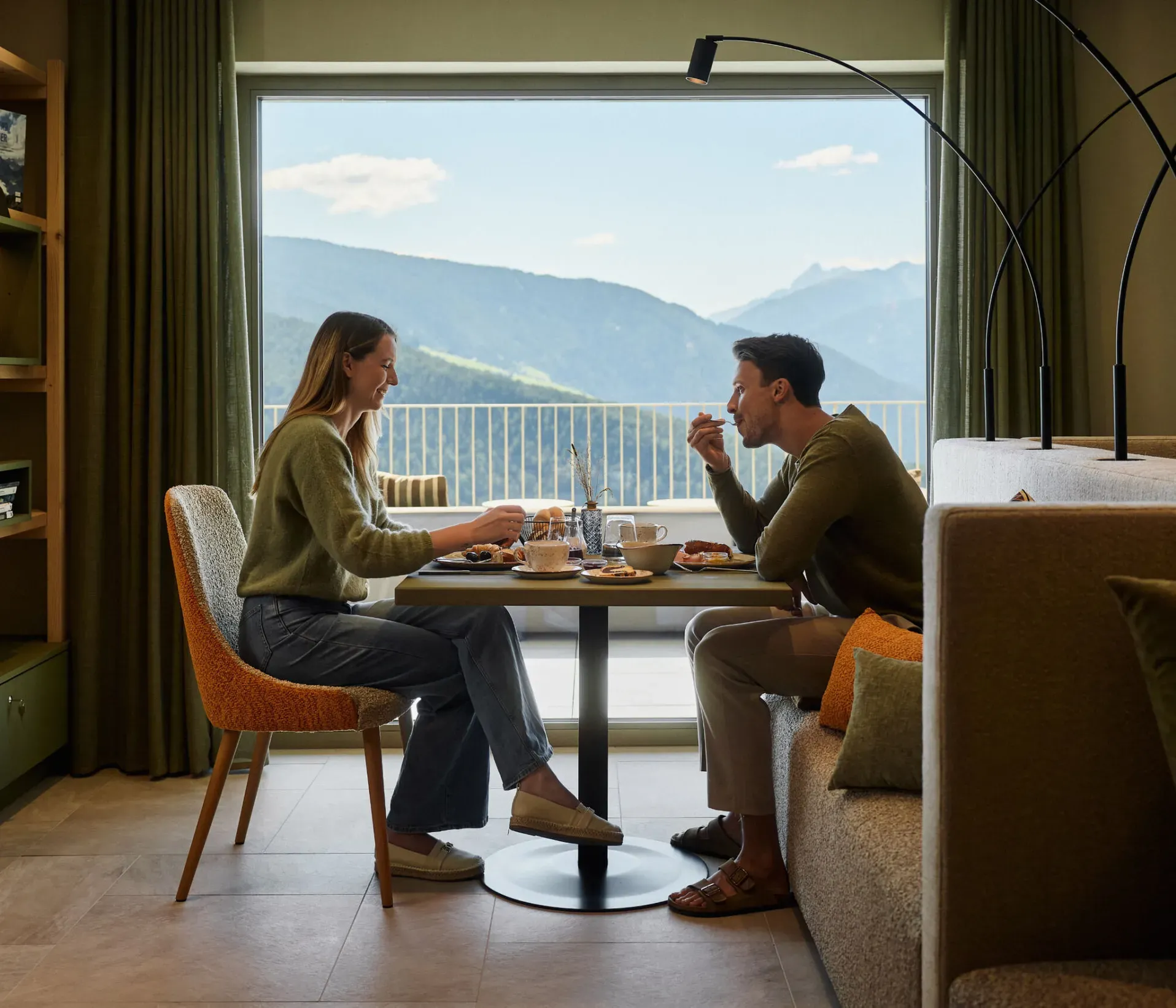 Una coppia si gode la colazione al tavolo dell’hotel AUI, con le montagne visibili sullo sfondo attraverso la finestra.