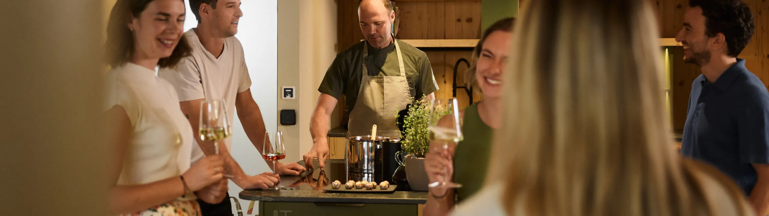 Gli ospiti si riuniscono attorno al bancone della cucina, gustando aperitivi e calici di vino durante un’esperienza culinaria all’Hotel AUI.