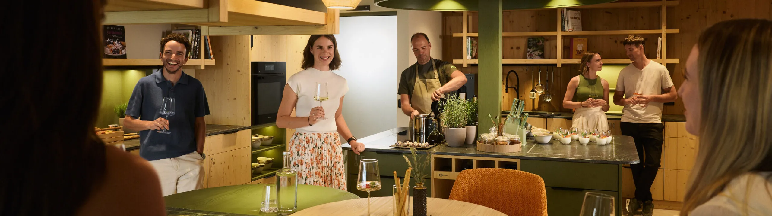 Ospiti e padroni di casa si riuniscono attorno a un tavolo da cucina nell’Hotel AUI, condividendo vino e conversazioni in un’atmosfera conviviale.