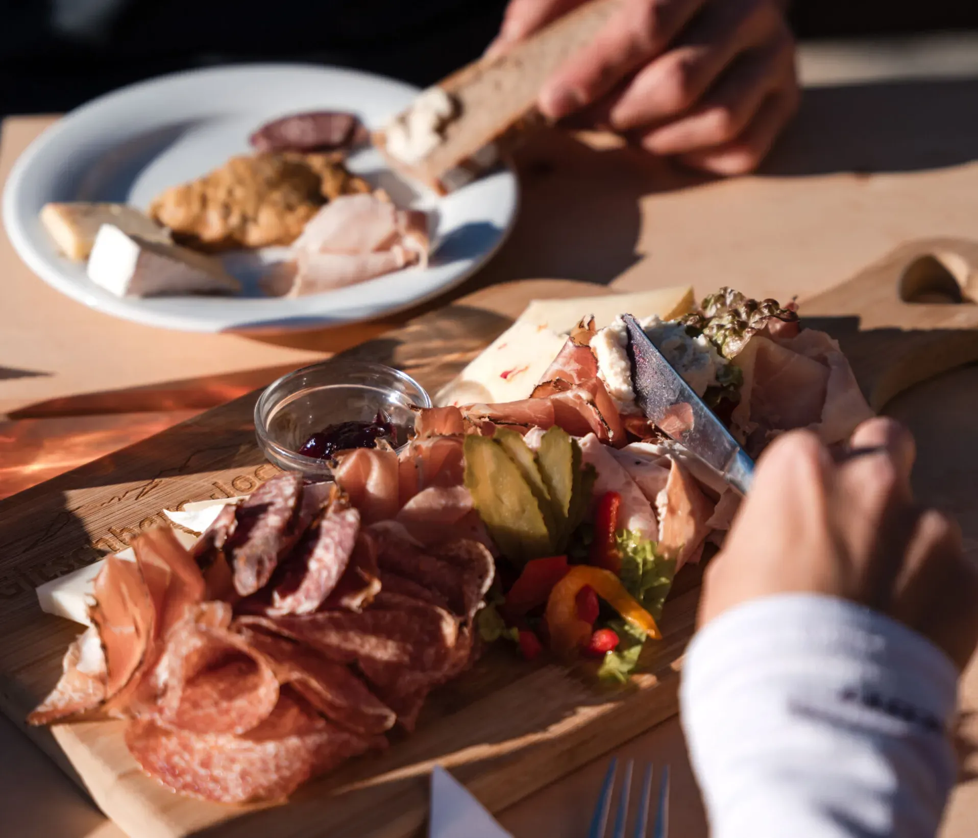 A wooden charcuterie board with assorted meats, pickles, and a small jam jar. Two people enjoy food, creating a cozy, casual dining atmosphere.
