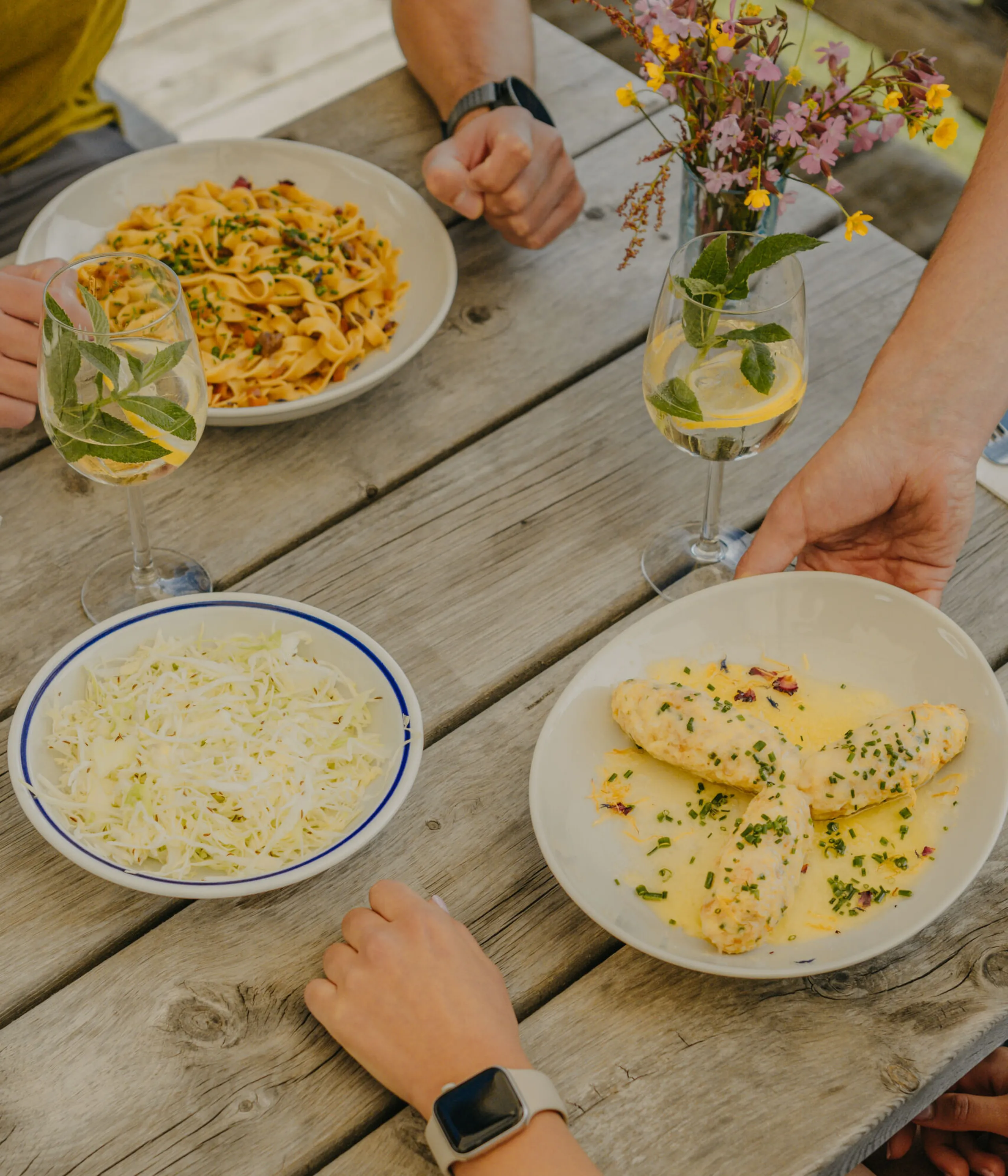 A casual outdoor meal on a wooden table featuring pasta, chicken in sauce, and salad. Two people dine, with drinks and a vase of flowers adding a cozy touch.