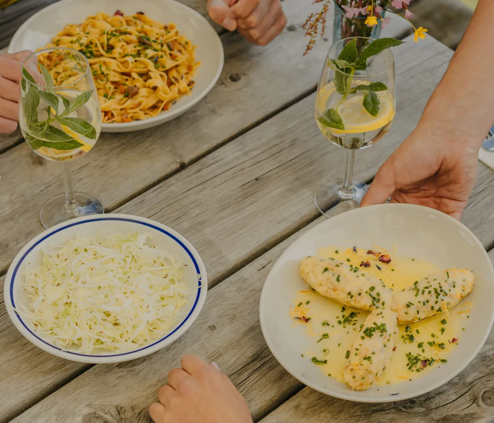 Ein ungezwungenes Essen im Freien auf einem Holztisch mit Pasta, Hühnchen in Soße und Salat. Zwei Personen essen zusammen, Getränke und eine Vase mit Blumen verleihen eine gemütliche Atmosphäre.