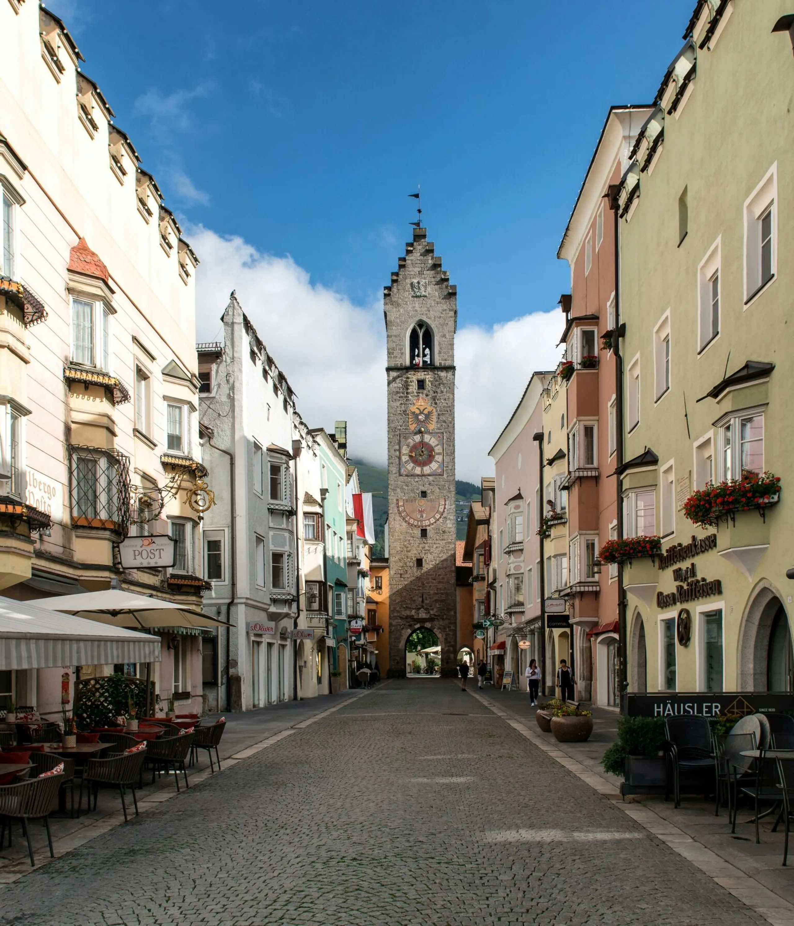 Una stretta strada acciottolata fiancheggiata da edifici colorati e caffè all’aperto conduce a un’alta torre dell’orologio ornamentale sotto un cielo azzurro e luminoso.