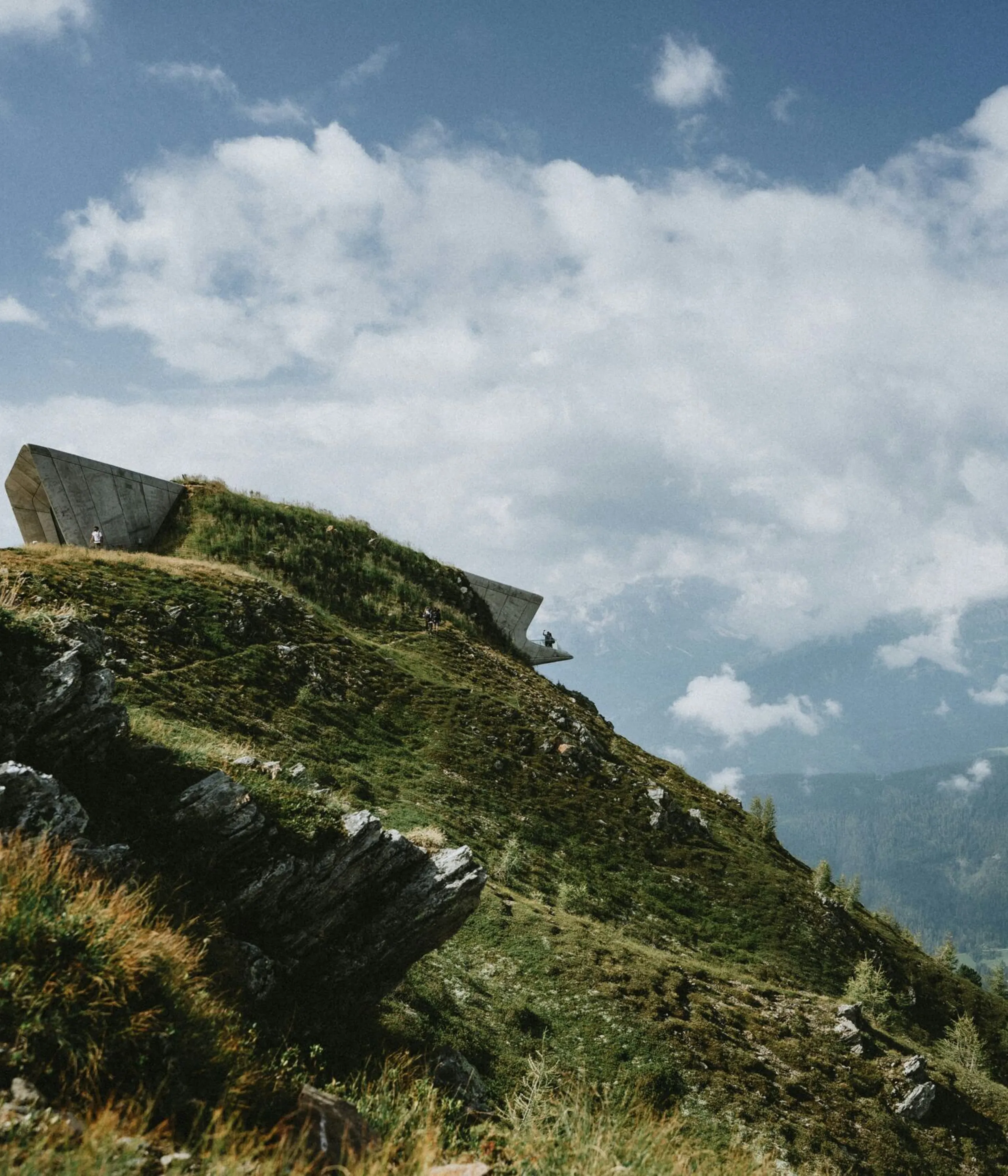 Un edificio moderno situato sulla cima di una collina erbosa sotto un cielo parzialmente nuvoloso, con montagne in lontananza sullo sfondo. Due piccole figure si trovano nelle vicinanze.