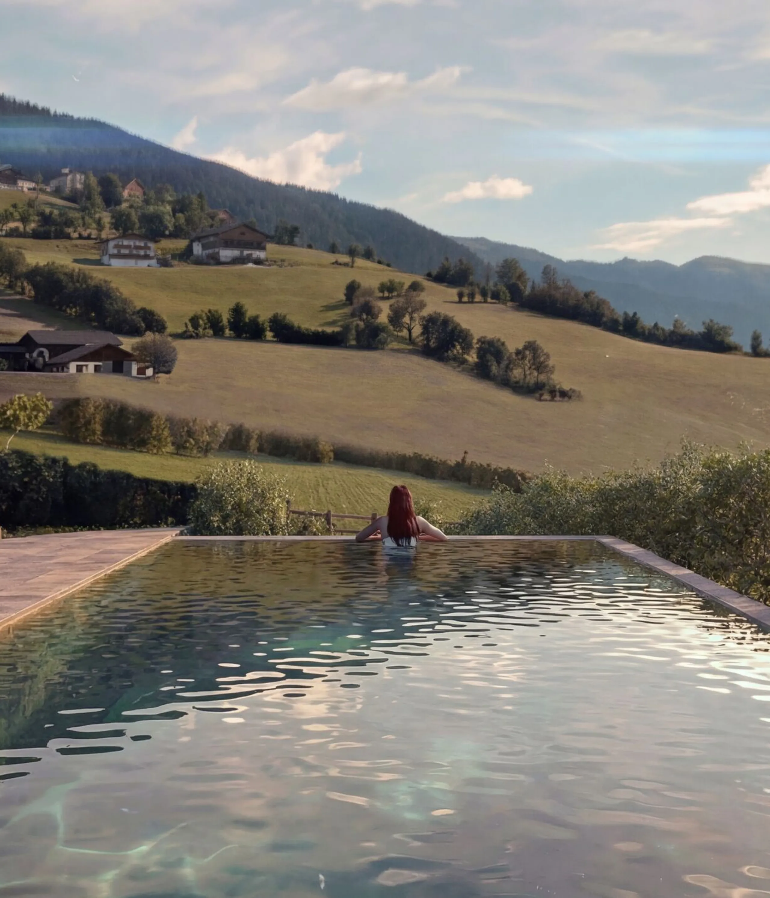 A person relaxes at the edge of an infinity pool, overlooking serene rolling hills and distant mountains under a partly cloudy sky, conveying tranquility.
