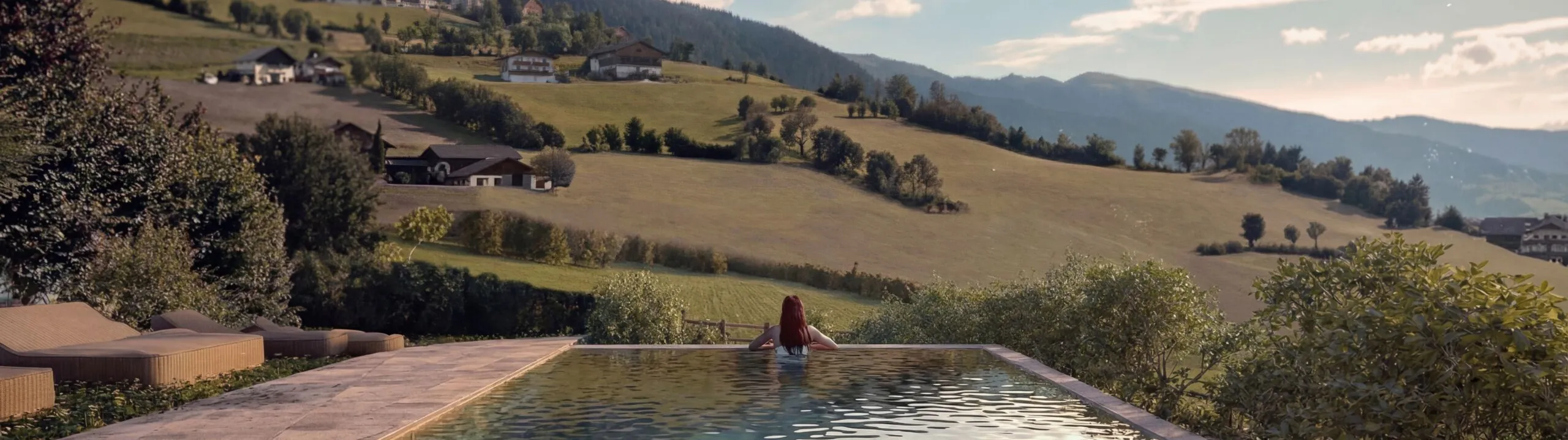 A person relaxes at the edge of an infinity pool, overlooking serene rolling hills and distant mountains under a partly cloudy sky, conveying tranquility.