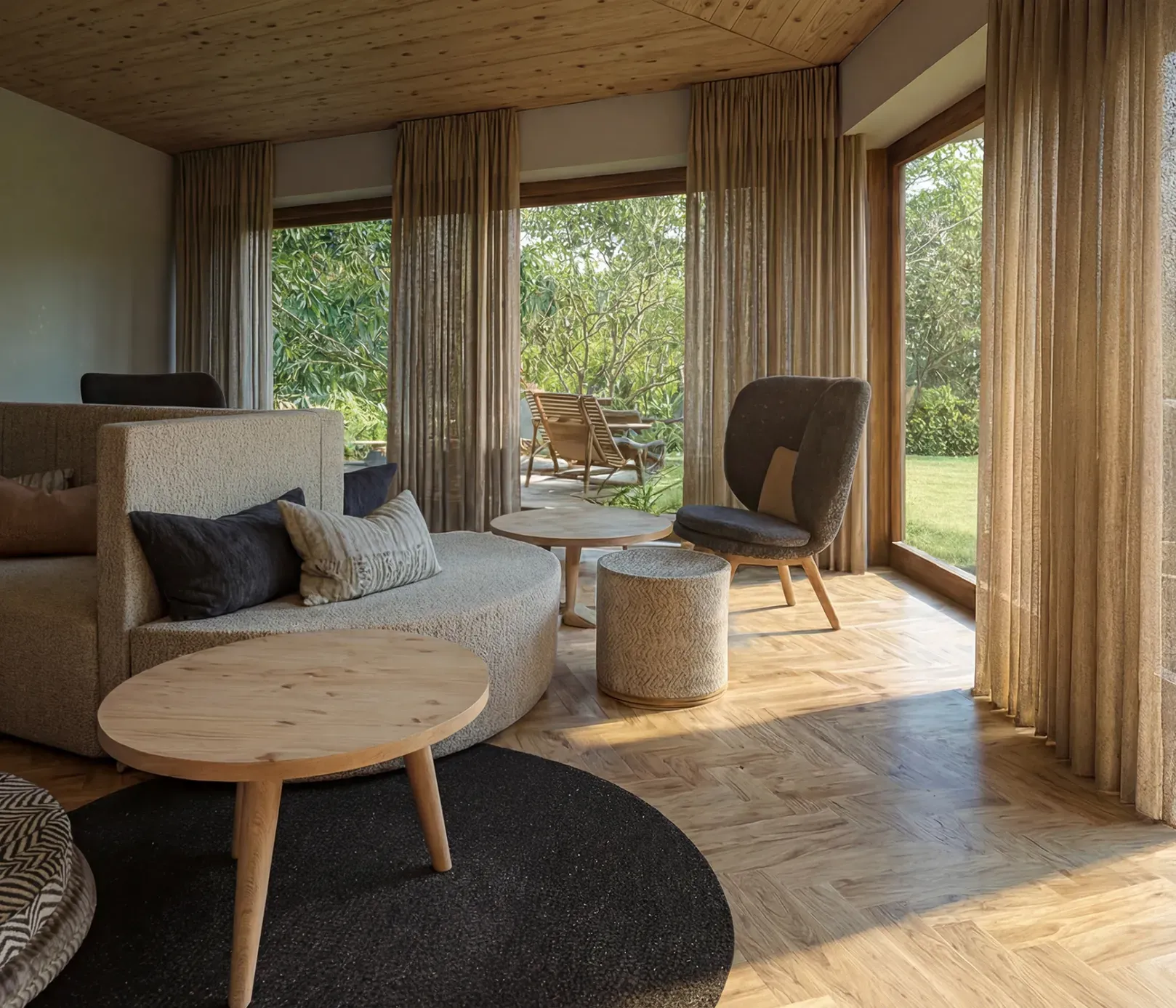 Stylish living room with large windows revealing a lush garden view. Cozy sofas and round wooden tables add warmth to the bright, modern space.