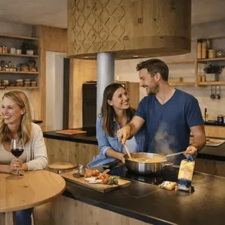 A cheerful group of four friends enjoy cooking in a warm, rustic kitchen. Two hold glasses of red wine, while another stirs pasta on the stove.