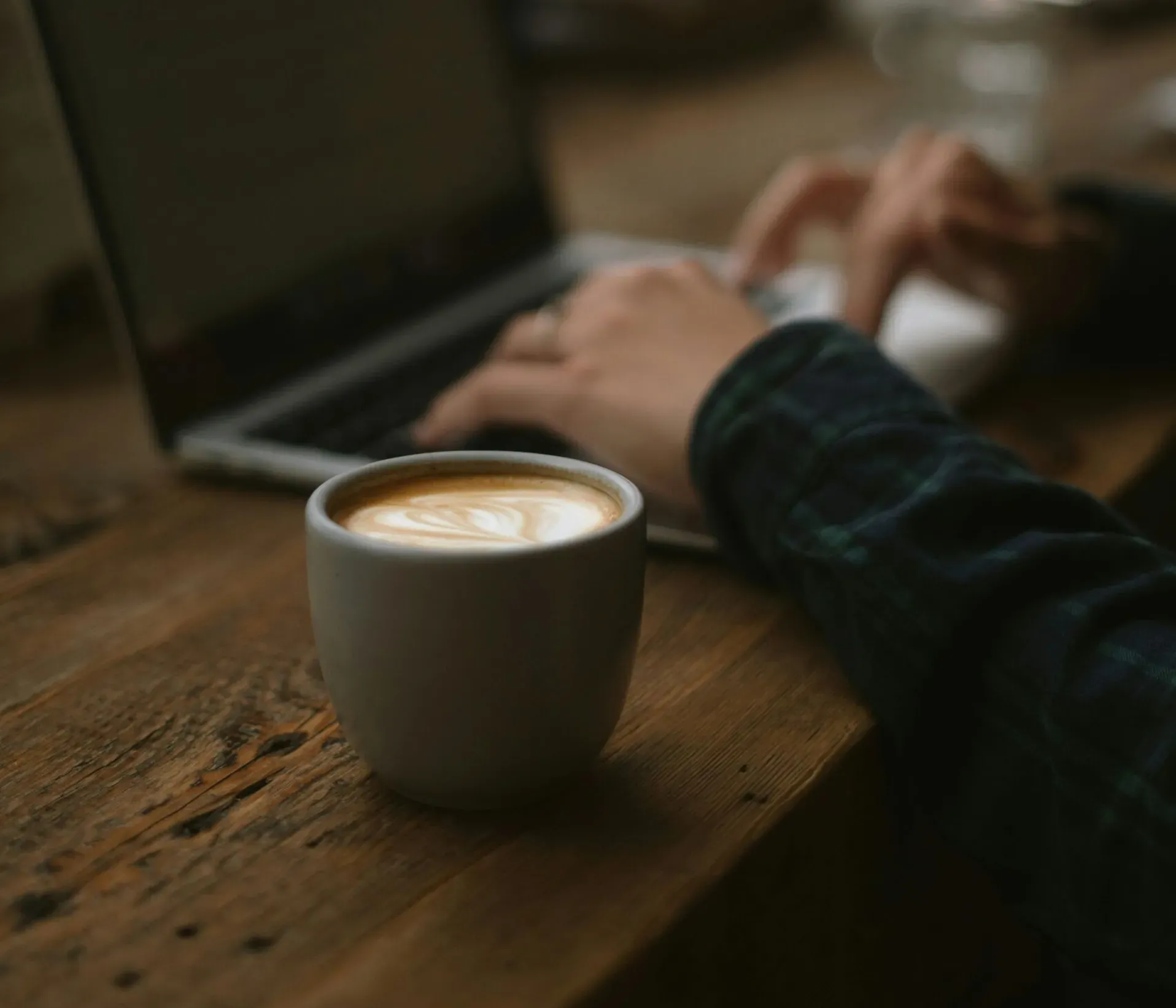 Una persona digita su un laptop a un tavolo di legno, con una tazza di caffè in primo piano. L’ambiente è accogliente e concentrato, suggerendo un tranquillo caffè.