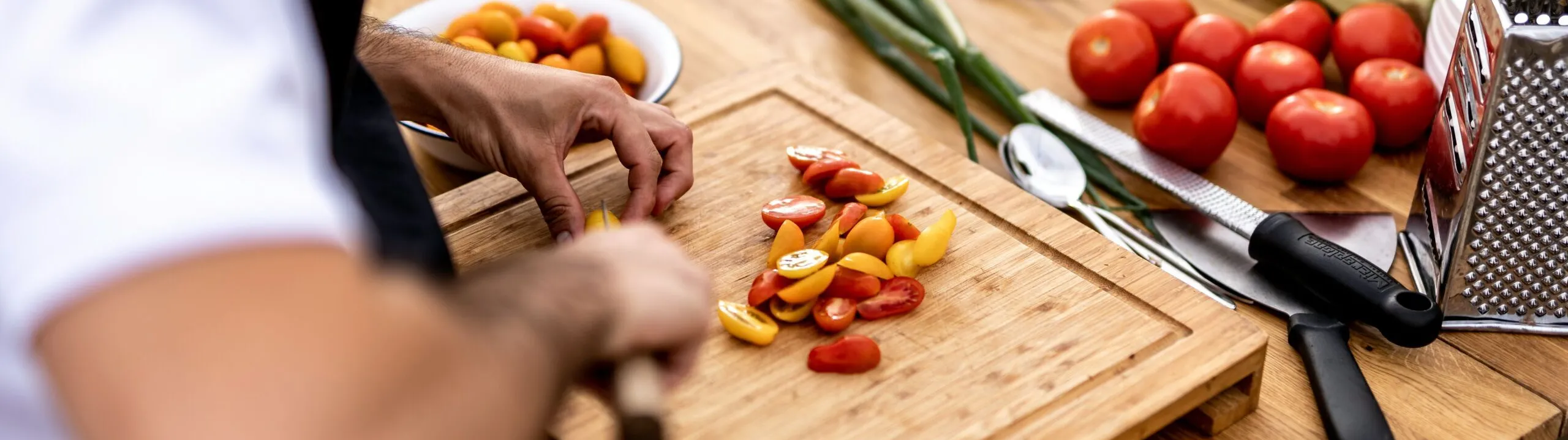 Person chopping cherry tomatoes on a wooden cutting board. Nearby are fresh vegetables, a knife, and a cheese grater on a wooden table.