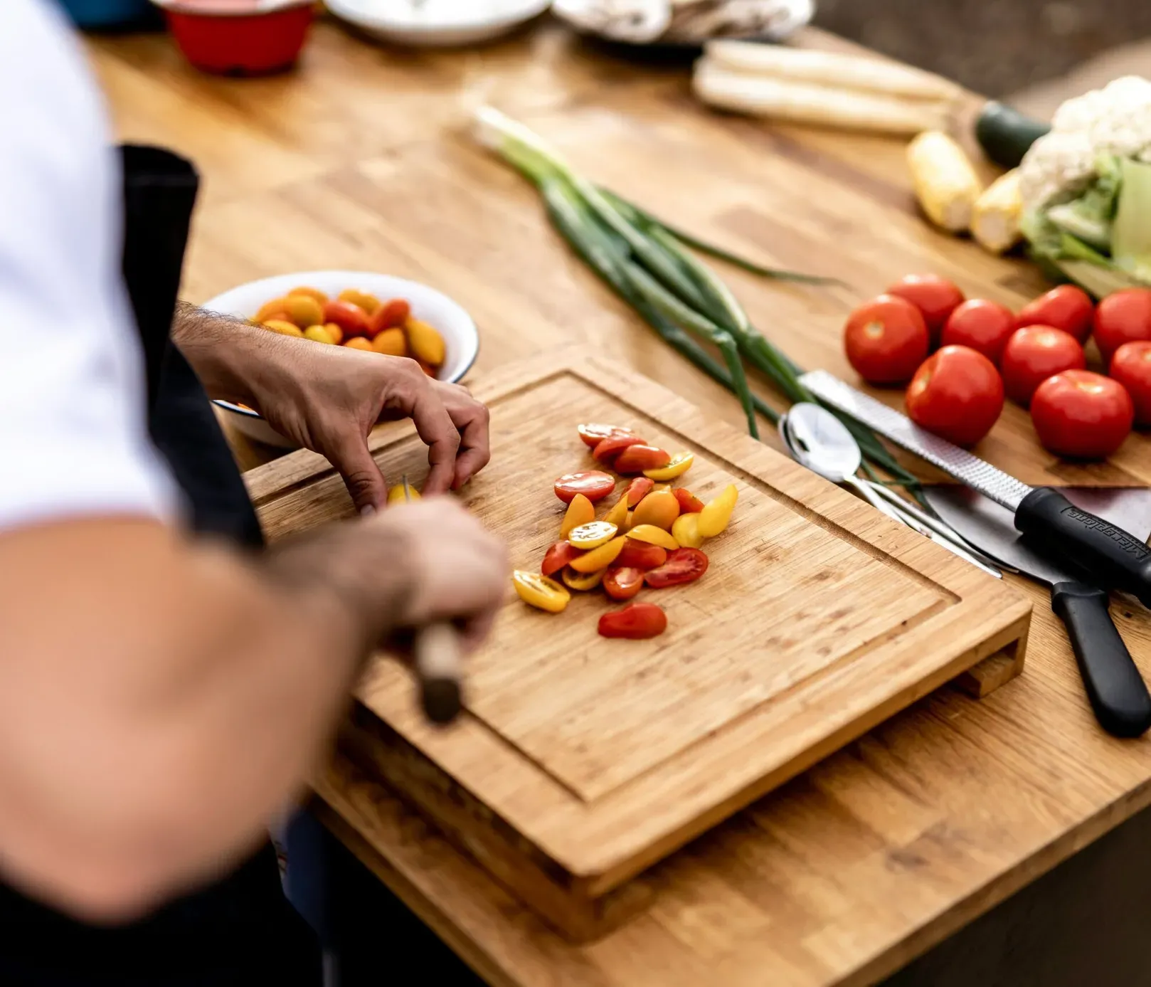Person schneidet Kirschtomaten auf einem Holzbrett. In der Nähe liegen frisches Gemüse, ein Messer und eine Reibe auf einem Holztisch.