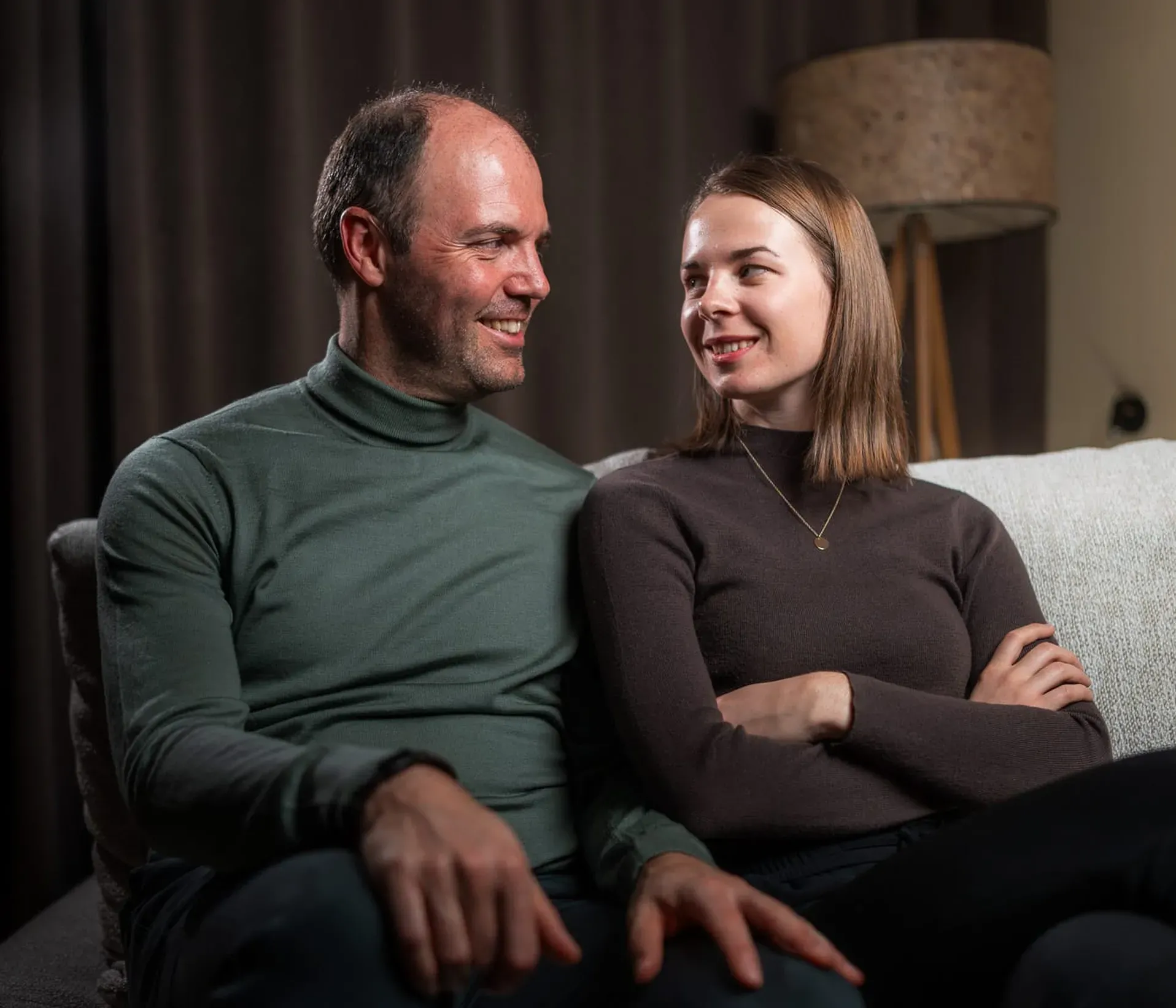 A man and a woman sit closely on a sofa, smiling warmly at each other, creating a cozy and affectionate atmosphere. A lamp and curtains are in the background.