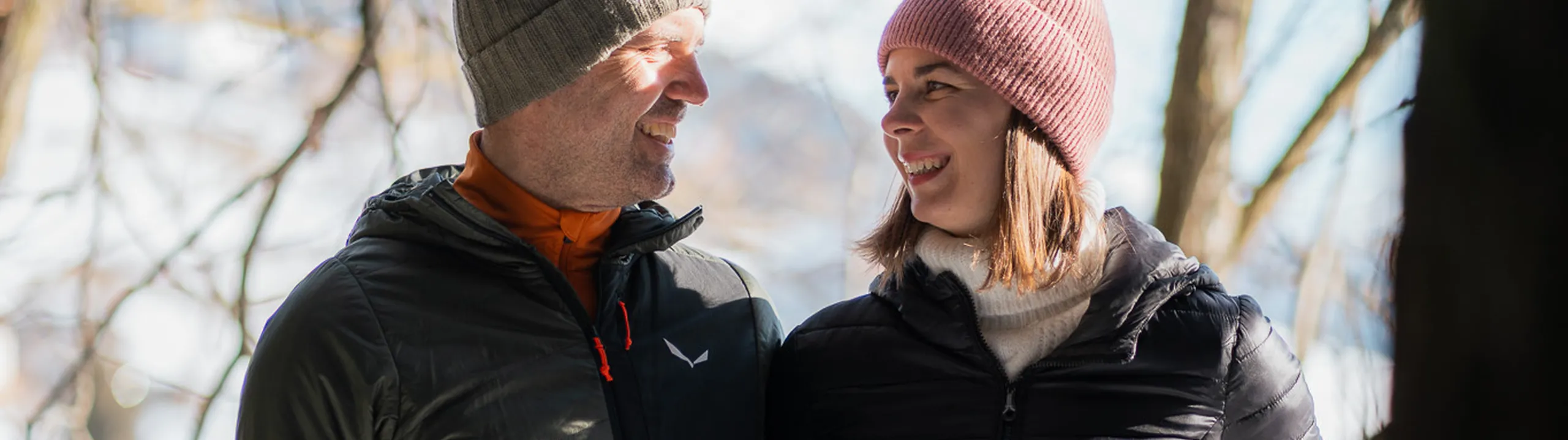 Ein Paar in Winterkleidung, mit Mützen und Jacken, steht lächelnd auf einem verschneiten Balkon im Wald und schaut sich an. Sonnenlicht fällt durch die kahlen Bäume.